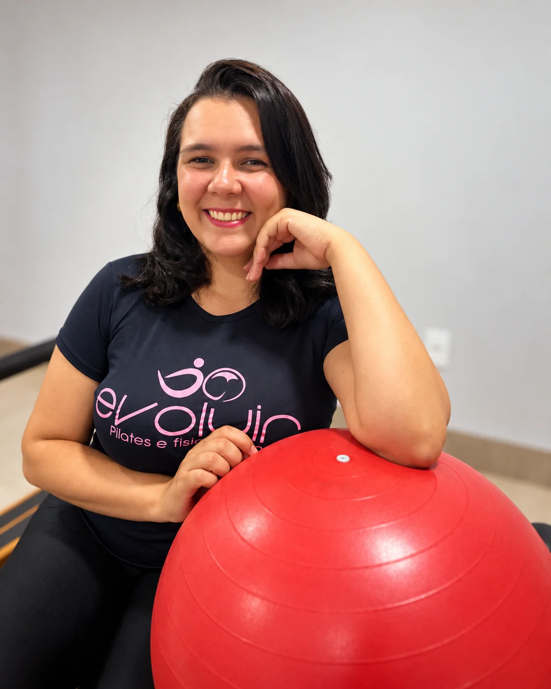 Paula Leylane Tavares Brito sorrindo no estúdio da Evoluir, apoiada em uma bola vermelha de Pilates.